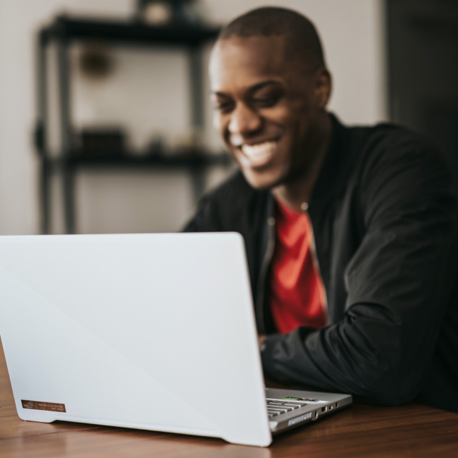 Un homme à l'expression joyeuse est assis à une table, absorbé par son travail sur un ordinateur portable.