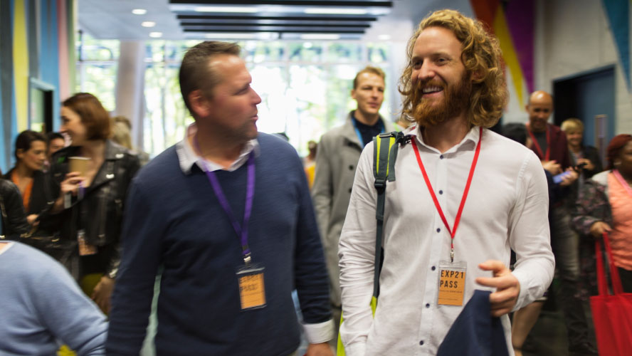 A bearded man walks down a hallway, carrying a backpack.