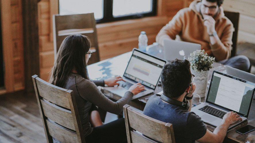 Trois personnes travaillant sur des ordinateurs portables à une table, absorbées par leurs tâches.