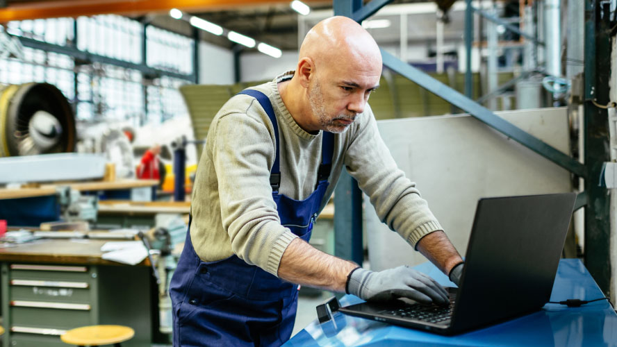 Un homme dans un environnement industriel, concentré sur son ordinateur portable, travaillant avec diligence sur des tâches.