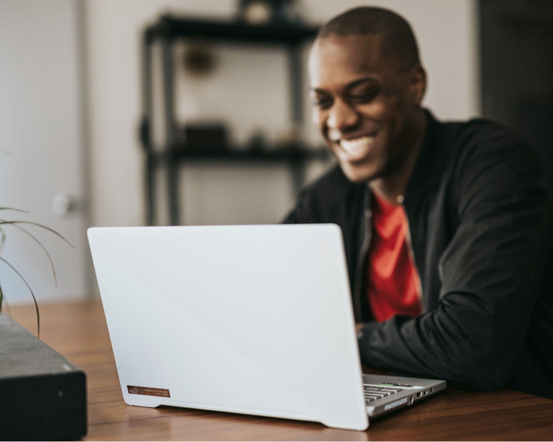 Un homme à l'expression joyeuse est assis à une table, absorbé par son travail sur un ordinateur portable.