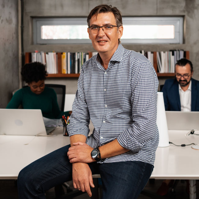 Un homme portant des lunettes est assis à un bureau avec deux ordinateurs portables, absorbé par son travail, faisant preuve de concentration et de productivité.