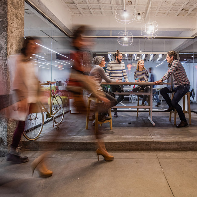 Group of colleagues discuss work at a table while others walk by